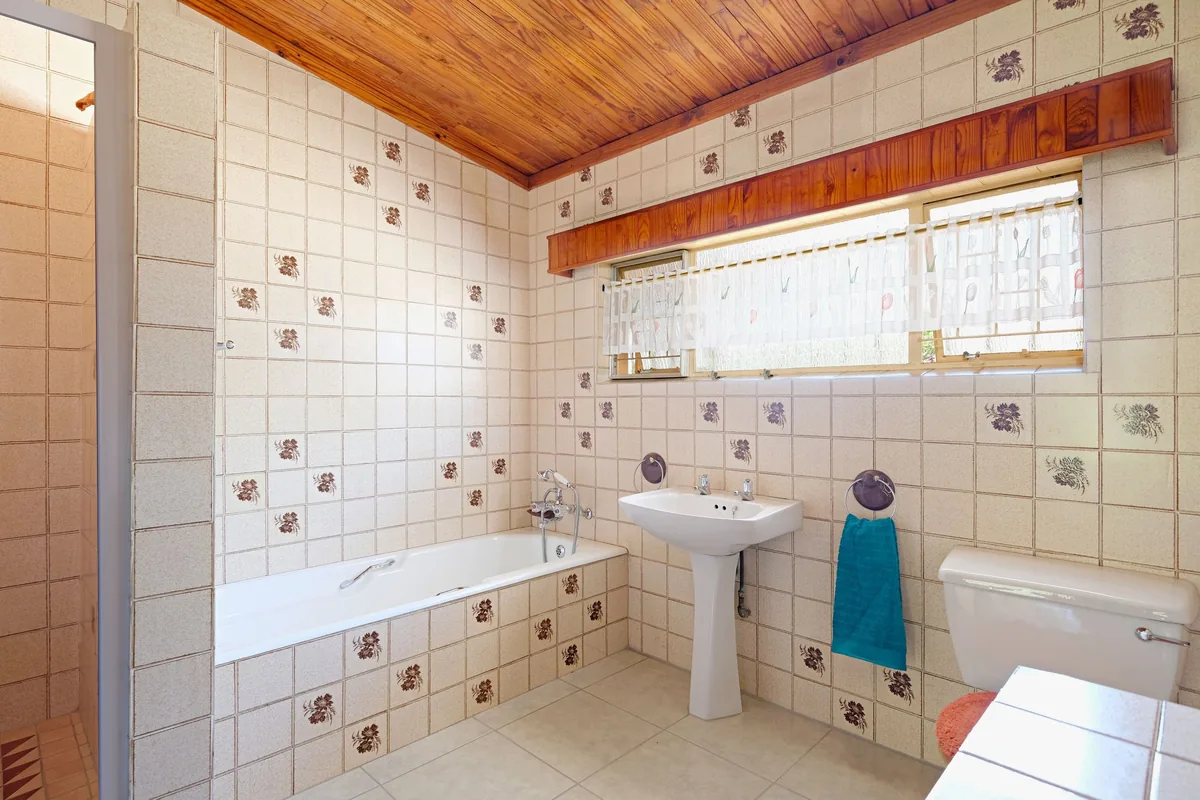Dated bathroom with floral wall tiles, pine ceiling, pedestal sink and a long window before AI redesign