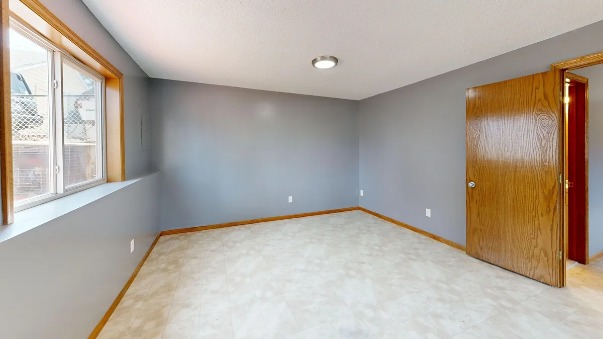 Empty basement room with grey walls, vinyl floor, a wide window and an oak door before AI redesign